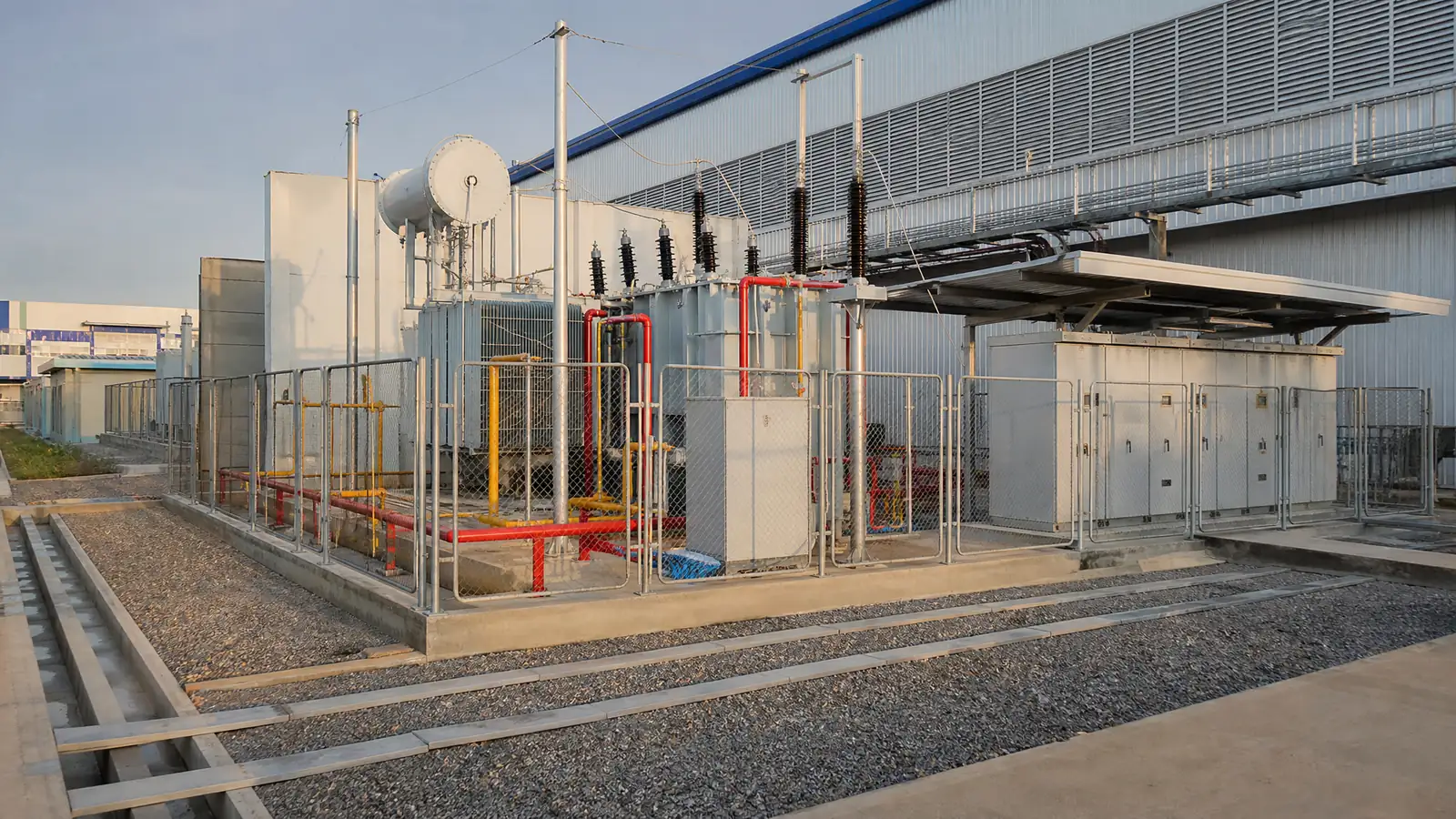 A transformer yard and large main switchboard beside a factory building, seen from a distance in afternoon light